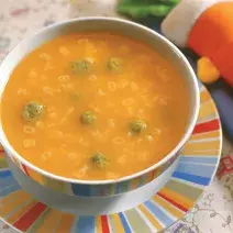 Fotografia em tons de laranja em uma mesa com uma toalha de folhas, um prato raso colorido, um prato fundo redondo colorido com a sopa divertida com bolinhas de espinafre.