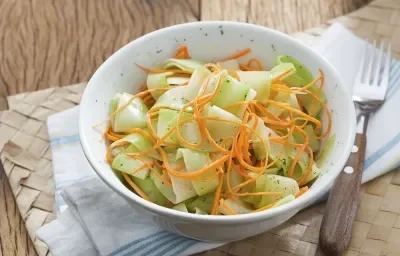 Fotografia em tons de verde em uma mesa de madeira com um pano branco com detalhes em azul e um prato fundo branco com a salada de chuchu dentro.