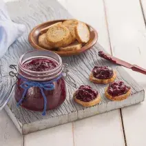 Fotografia em tons de azul em uma bancada de madeira clara, uma tábua de madeira, um paninho azul, um pratinho com algumas torradas integrais e um pote hermético de vidro com a geleia de frutas vermelhas dentro.