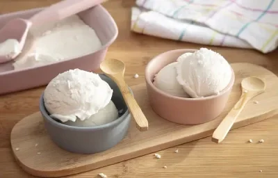 Foto em tons claros da receita de sorvete de coco cremoso caseiro servida em duas porções em potinhos azul e rosa com colheres de madeira sobre uma tábua de madeira com a receita maior ao fundo Foto em tons claros da receita de sorvete de coco cremoso caseiro servida em duas porções em potinhos azul e rosa com colheres de madeira sobre uma tábua de madeira com a receita maior ao fundo