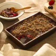 Foto em tons de marrom da receita de doce de aveia com frutas vermelhas servida em uma forma de madeira marrom escura com dois pratos fundos brancos ao lado com porções do doce