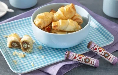 Foto da receita de folhado de baton algodão doce servida em algumas porções dentro de um bowl de porcelana azul claro em cima de uma base quadriculada com unidades de baton ao lado