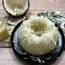 Foto da receita de Cuscuz de Tapioca. Observa-se um cuscuz decorado com coco ralado com um pote transparente com leite moça ao lado.