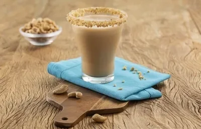 Foto da Receita de Cappuccino Gelado de Caju. Observa-se um copo transparente de vidro decorado com castanha de caju no topo sobre um guardanapo azul e uma pequena tábua de madeira. Atrás, tem um potinho com castanhas de caju.