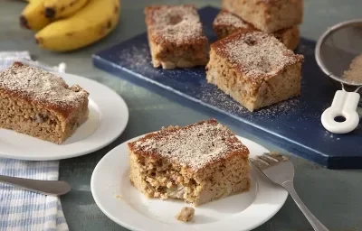 Foto da receita de bolo de banana com farinha láctea aveia servida em duas porções dispostas sobre pratos de porcelana brancos com uma régua azul atrás com mais pedaços do bolo em cima. Ao fundo ainda há duas bananas e uma peneira com canela em pó