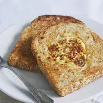 Fotografia em tons de bege em uma mesa de madeira clara com um prato branco raso e duas fatias de pão integral com a omelete dentro dele.