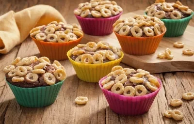 Fotografia em tons de laranja, verde, amarelo e rosa em uma bancada de madeira clara, um paninho amarelo, uma tábua de madeira, e várias forminhas de doces coloridas com o cupcake de chocolate cobertos com cheerios.