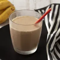 Fotografia em tons de branco e preto de uma bancada de madeira branca com um prato preto, sobre ele um copo de vidro com a bebida cremosa e um canudo vermelho. Ao lado um paninho listrado branco e preto e um cacho de banana.