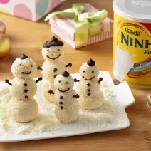 Fotografia em tons de marrom de uma bancada de madeira com uma tábua branca, sobre ela quatro bonecos de neve. Ao lado uma colher, uma lata de NINHO. Ao fundo bolinhas natalinas e caixinhas de presente.