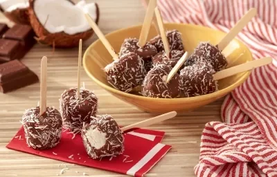 Fotografia em tons de marrom em uma bancada de madeira de cor marrom. Ao centro, um recipiente contendo os picolés. Ao lado, há alguns picolés em cima de um pano vermelho. Ao fundo, um pano vermelho listrado e um coco e chocolates espalhados.