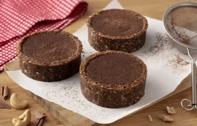 Fotografia em tons de marrom e vermelho de uma bancada de madeira com uma tábua de madeira e uma folha de papel manteiga, sobre ele três tortinhas de ganache. Ao fundo um paninho xadrez vermelho e branco e uma peneira. A frente castanhas-de-caju.