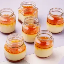 fotografia em tons de branco e laranja vista de cima, contém 6 potinhos transparentes com pudim de dentro e cobertura de caramelo por cima