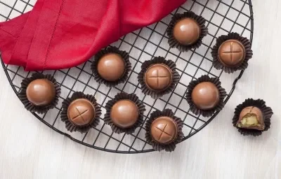 Fotografia em tons de chocolate, branco e vermelho, ao centro vários bombons de chocolate em forminhas de papel marrom sobre grelha redonda, ao lado guardanapo vermelho e bombom aberto, tudo sobre bancada de madeira clara.