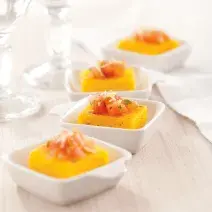 Fotografia em tons de laranja em uma bancada de madeira clara, um pano bege, quatro potinhos brancos pequenos com a polenta com tomate dentro de cada um.