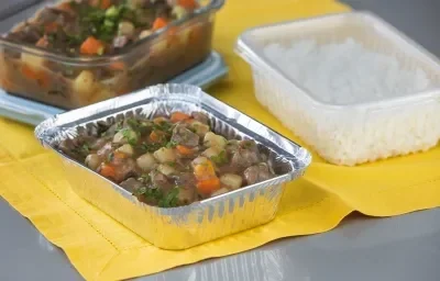 Foto da receita de picadinho de carne, servido em um recipiente de alumínio, sobre uma toalha amarela e acompanhado de arroz num pote de plástico Foto da receita de picadinho de carne, servido em um recipiente de alumínio, sobre uma toalha amarela e acompanhado de arroz num pote de plástico
