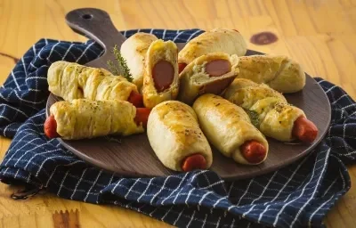 Foto aproximada de uma bancada sobre a qual está um pano azul quadriculado e uma tábua com a receita de enroladinho de salsicha nela. Foto aproximada de uma bancada sobre a qual está um pano azul quadriculado e uma tábua com a receita de enroladinho de salsicha nela.