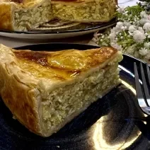 Foto da receita de Torta de Cebola e Ovos. Observa-se um pedaço grande de torta em um prato preto com decoração de flores brancas.