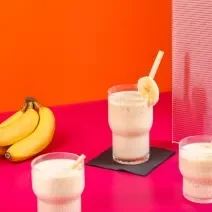 Fotografia de três copos de vidro com vitamina de banana e goiaba, e em cada copo, tem um canudo e uma rodela de banana decorando-o. Ao lado esquerdo, um cacho com quatro bananas, sobre uma mesa rosa com fundo laranja.
