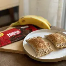 Fotografia em tons de marrom em uma bancada de madeira com uma tábua de madeira e ao centro, um prato redondo com dois pasteis. Ao lado, um tablete de chocolate e um cacho de bananas ao fundo.