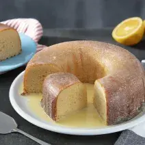 Fotografia em tons de amarelo em uma bancada de madeira cor preta. Ao centro, um prato branco contendo o bolo com uma espátula ao lado. Ao fundo, há um pires azul com uma fatia do bolo e uma laranja partida ao meio.
