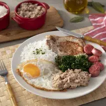 Fotografia em tons de vermelho em uma bancada de madeira de cor marrom. Ao centro, um prato branco contendo o virado à paulista. Ao lado, há um garfo, uma faca e um pano vermelho listrado. Ao fundo, há um pouco de arroz e tutu de feijão.