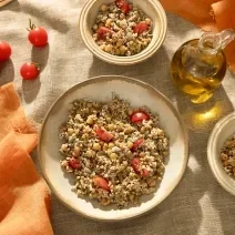 Foto da receita de salada de grãos com aveia servida em pratos fundos de cerâmica clara com tomates cereja e um vidro de azeite ao lado além de um paninho laranja