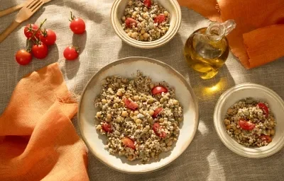 Foto da receita de salada de grãos com aveia servida em pratos fundos de cerâmica clara com tomates cereja e um vidro de azeite ao lado além de um paninho laranja