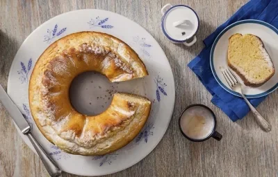 Fotografia em tons de cinza, branco e azul de uma bancada cinza e um prato banco com detalhes azuis, sobre ele o bolo de milho. Ao lado duas xícaras de café com leite e um paninho azul com um prato branco de bordas azuis com uma fatia do bolo e um garfo.