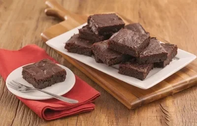 Fotografia em tons de marrom e vermelho de uma bancada de madeira com uma tábua de madeira, sobre ele um papel manteiga com pedaços de brownie. Ao fundo um prato com um pedaço de brownie e um garfo. Fotografia em tons de marrom e vermelho de uma bancada de madeira com uma tábua de madeira, sobre ele um papel manteiga com pedaços de brownie. Ao fundo um prato com um pedaço de brownie e um garfo.