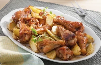 Fotografia em tons de amarelo em uma bancada de madeira com um pano branco com em detalhes amarelos, com um recipiente branco oval com as coxinhas da asa assadas com batata crocantes.