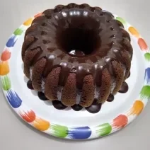 Foto em tons de marrom da receita de bolo de café servida com uma cobertura de chocolate por cima e sobre um prato branco com a borda colorida