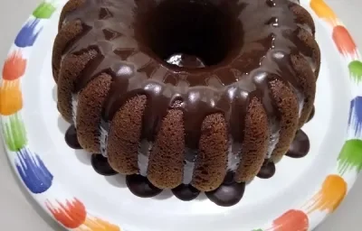 Foto em tons de marrom da receita de bolo de café servida com uma cobertura de chocolate por cima e sobre um prato branco com a borda colorida