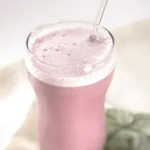 Fotografia em tons de lilás e branco. Um copo transparente contém suco de uva com espuma por cima e um canudo transparente.