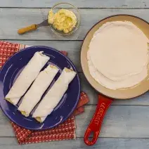 Fotografia em tons de vermelho e azul em uma bancada de madeira com um pano vermelho, um prato azul com três beiju de manteiga, uma frigideira grande vermelha com a massa de beiju dentro dela.