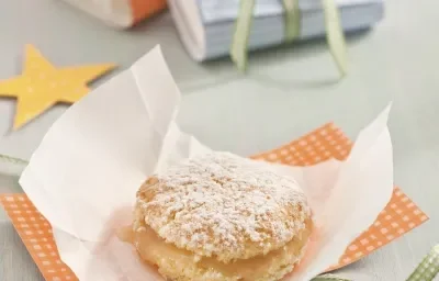 Fotografia em tons de laranja, amarelo e azul em uma bancada cinza. Um pedaço de papel laranja com bolinhas e em cima dele um bem-casado, ao lado estrelas de papel na cor azul e amarela. Ao fundo, três bem-casados embrulhados em papeis coloridos.