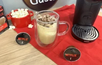 Fotografia em tons de vermelho em uma bancada de madeira clara, um pano vermelho, uma xícara de vidro com a bebida de café com sorvete dentro. Ao lado, um pote vermelho de pipoca e uma máquina dolce gusto. Fotografia em tons de vermelho em uma bancada de madeira clara, um pano vermelho, uma xícara de vidro com a bebida de café com sorvete dentro. Ao lado, um pote vermelho de pipoca e uma máquina dolce gusto.