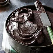 Fotografia em tons de marrom, em uma bancada de madeira de cor cinza. Ao centro, uma forma redonda contendo o bolo com uma espátula em cima.