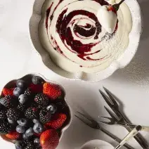 Fotografia em tons de branco, em uma mesa de madeira de cor branca. Ao centro, um recipiente contendo o fondue e ao lado um outro recipiente contendo as frutas. Ao fundo, alguns garfos.