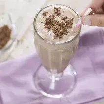 Fotografia em tons e branco e roxo de uma bancada branca com um paninho roxo, sobre ele uma taça de vidro com milk shake e um canudo. Ao lado um recipiente de vidro com raspas de chocolate.