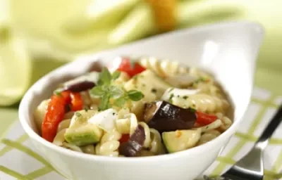 Fotografia em tons de verde em uma mesa de madeira verde com um guardanapo de papel quadriculado em branco e verde e um potinho branco com a salada mediterrânea dentro.