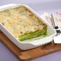 Fotografia em tons de branco e verde, com refratário branco contendo uma lasanha de espinafre gratinada, sobre uma tábua de cozinha com uma espátula de servir sobre um guardanapo branco com bolinhas vermelhas em uma bancada lilás.