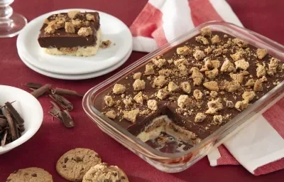 Fotografia de um pavê de chocolate com biscoito em uma travessa retangular transparente, com pedaços de biscoitos polvilhados por cima. Ao fundo uma toalha de mesa vermelha, com um prato de sobremesa branco servind um pedaço do pavê.