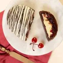 Foto da receita de Ovo Floresta Branca, cortado em um fatia, visto de cima, servido em um prato branco redondo, decorado com duas cerejas com cabo e uma faca dourada sobre um tecido vermelho