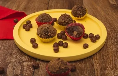 Fotografia em tons de amarelo em uma bancada de madeira escura, um pano vermelho, um prato amarelo redondo raso com brigadeiros de Nescau e recheados com Nescau Cereal.