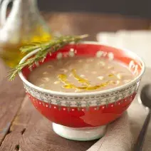 Fotografia em tons de vermelho em uma mesa de madeira com um recipiente redondo e fundo vermelho com a sopa de feijão dentro. Ao lado, um guardanapo de pano bege com uma colher apoiada nele e ao fundo, um potinho de azeite.