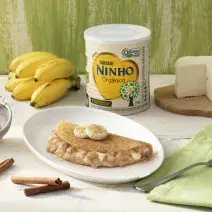 Fotografia em tons de branco e verde de uma bancada branca com um prato branco redondo com uma panqueca. Ao lado um paninho verde com um garfo, paus de canela, uma peneira, um cacho de banana, um queijo branco e uma lata de Ninho Orgânico.