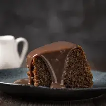 fotografia em tons de preto e marrom tirada de uma fatia de bolo de chocolate com calda por cima, e ao fundo um jarra branca