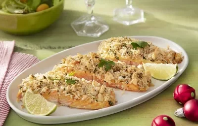 Foto da receita de salmão com crosta de parmesão sem glúten servida em três porções sobre uma base circular de porcelana em cima de uma mesa verde com um paninho quadriculado vermelho ao lado