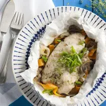 Fotografia em tons de azul em uma mesa de madeira azul com um prato branco fundo com detalhes azuis e o filé de peixe com legumes dentro dele.