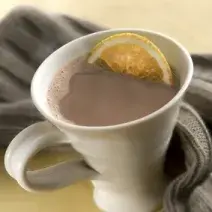 Fotografia em tons de cinza em uma bancada de madeira clara, um pano cinza escuro, uma xícara branca com o chocolate quente com uma rodela de laranja dentro.